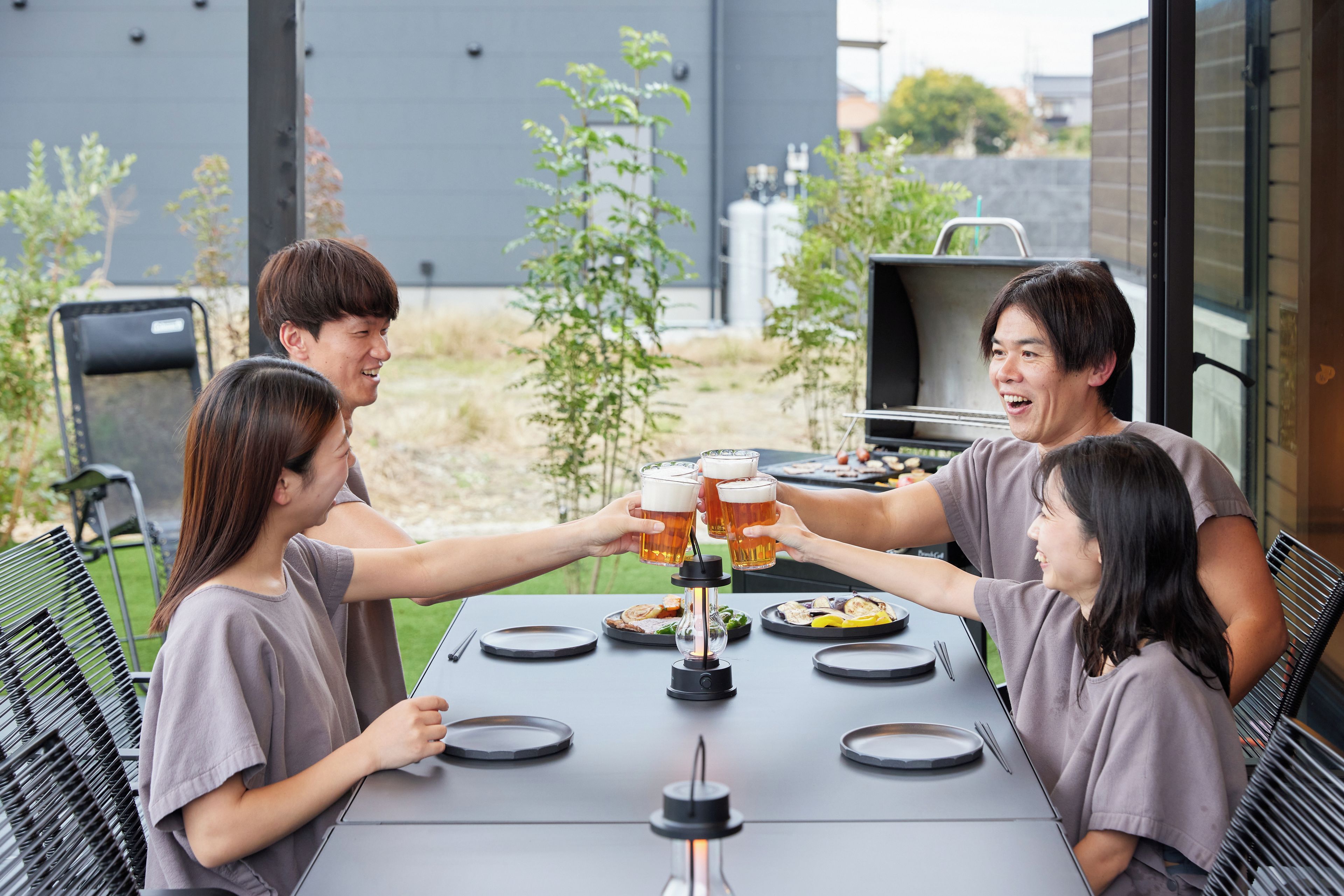 Un groupe de personnes trinquant avec des verres de bière autour d’une table où se trouvent un barbecue et des assiettes de nourriture.