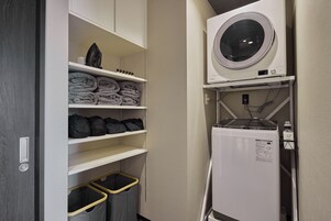 A laundry room with a front-loading washing machine, shelves with folded towels, and a trash bin.