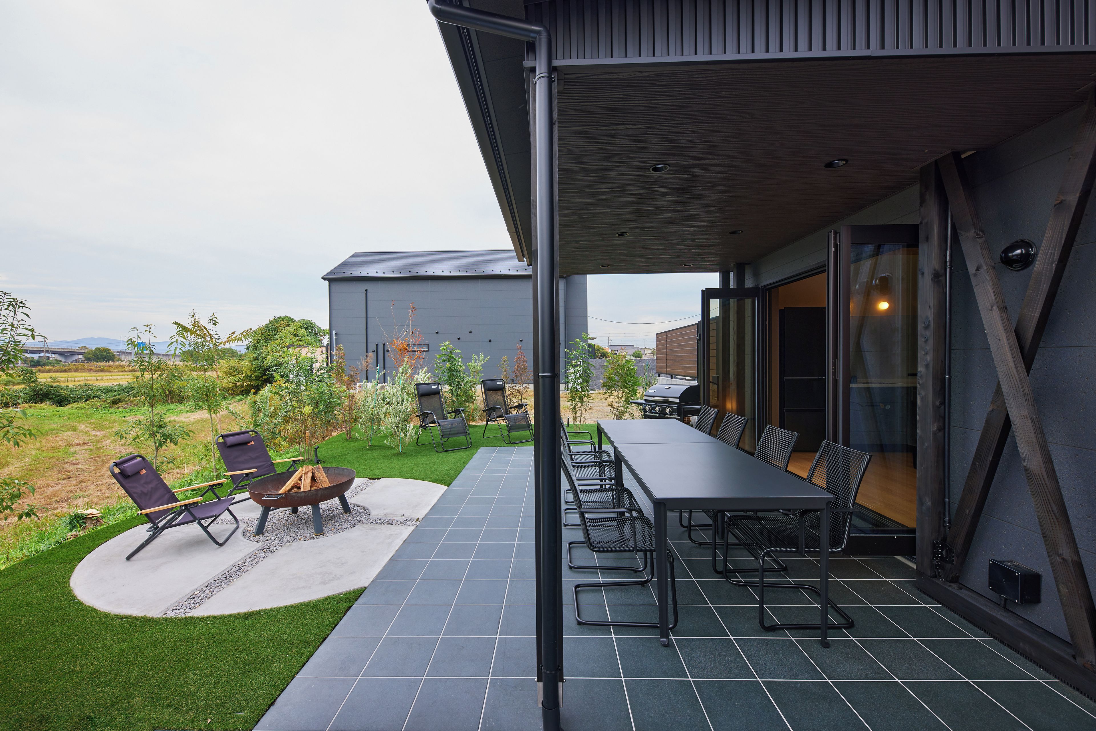 Un patio exterior con mesa y sillas, una chimenea y vistas a un jardín y un edificio al fondo.