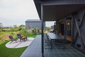 Un patio exterior con mesa y sillas, una chimenea y vistas a un jardín y un edificio al fondo.