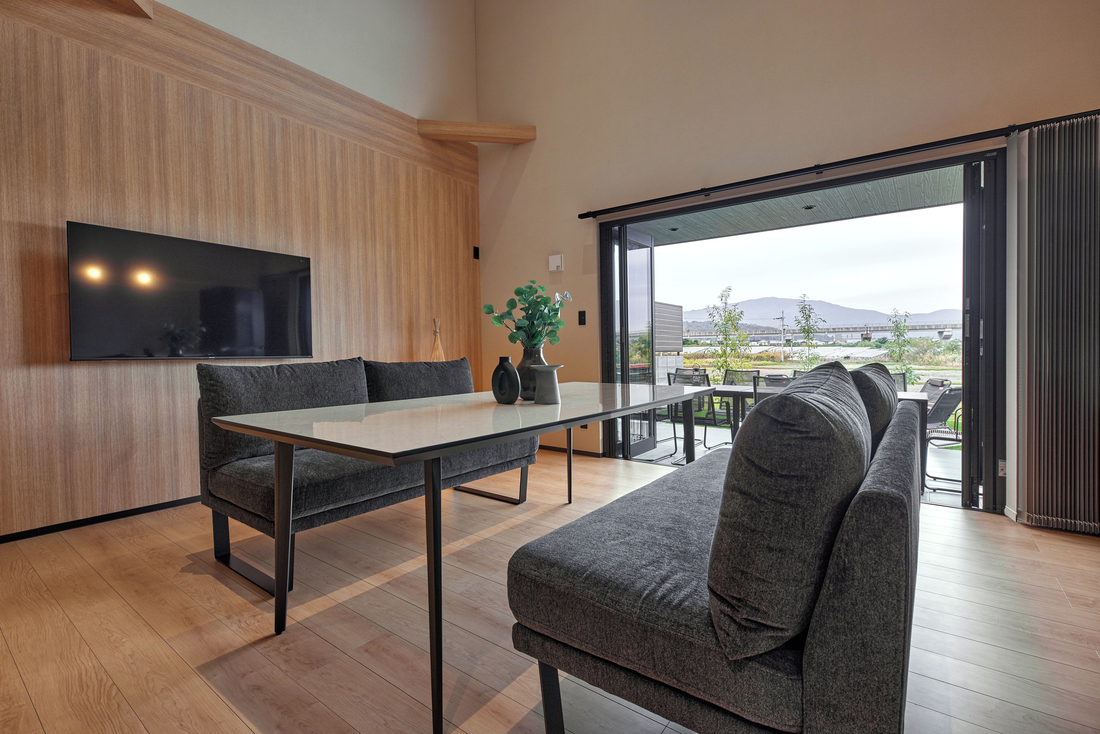 Un intérieur moderne avec un mur d’accent en bois, un téléviseur à écran plat, une table en verre, des canapés gris et une vue sur un pont et des montagnes, visible à travers de grandes portes vitrées.