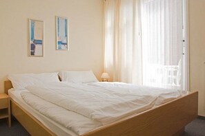 Ein Einzelbett mit Holzgestell, ein Nachttisch und ein Fenster mit durchsichtigen Vorhängen.