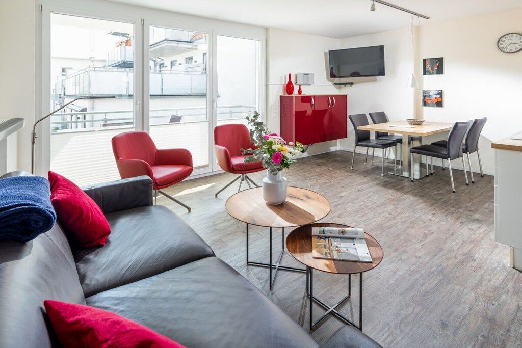 A modern living room with a grey sofa, red chairs, a wooden coffee table, and a dining area with a red cabinet and a TV.