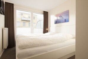 A hotel room with a bed, a window with curtains, a radiator, and a painting on the wall. - Holiday apartment in Norderney with Wi-Fi (Norderney)