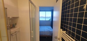 Ein schmales Badezimmer mit Dusche, einer kleinen Ablage für Toilettenartikel und Blick auf ein Bett durch eine offene Tür.