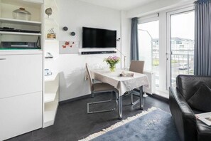 A modern living room with a flat-screen TV, a dining table with a vase of flowers, a sofa, and a view of the outside. - Holiday apartment in Norderney with sauna (Norderney)