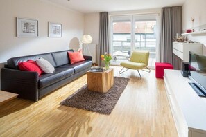 Ein modernes Wohnzimmer mit einem schwarzen Sofa, einem Holztisch, einem gelben Sessel und einem roten Hocker. Es gibt ein großes Fenster mit Blick auf Gebäude.