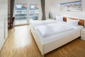 A hotel room with a double bed, a desk with a chair, a radiator, and a view of the outside balcony. - Holiday apartment in Norderney with Wi-Fi (Norderney)