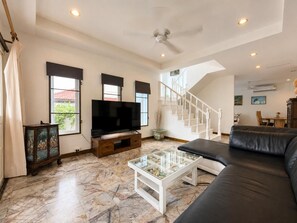 A spacious living room with a large flat-screen TV, a black leather sofa, a glass coffee table, and a staircase leading to an upper level. - Charming Thai Mountain Villa (MVV) (Kathu)