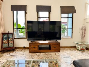 A living room with a flat-screen TV on a wooden stand, large windows with dark blinds, a traditional decorative cabinet, and a glass table with pebbles. - Charming Thai Mountain Villa (MVV) (Kathu)