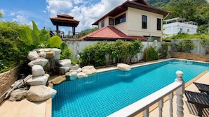 Una piscina con borde de azulejos, rodeada de rocas y follaje, con una casa y un pabellón al fondo.