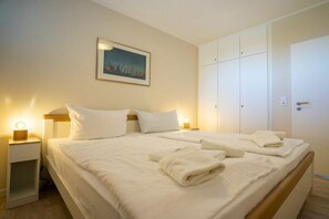 Una cama doble con ropa de cama blanca, dos mesitas de noche con lámparas, un armario y un cuadro en la pared.