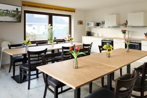 A dining area with a long table and chairs, kitchen area with appliances, and a window with a view. - Quarter 6 Apartment 5 (Büsum)