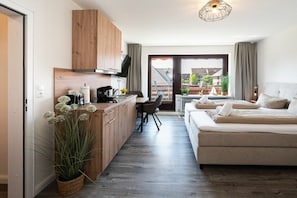 A modern kitchen with wooden cabinets, a dining area with a table and chairs, and a large bed with a view to the outside. - Quarter 6 Apartment 5 (Büsum)