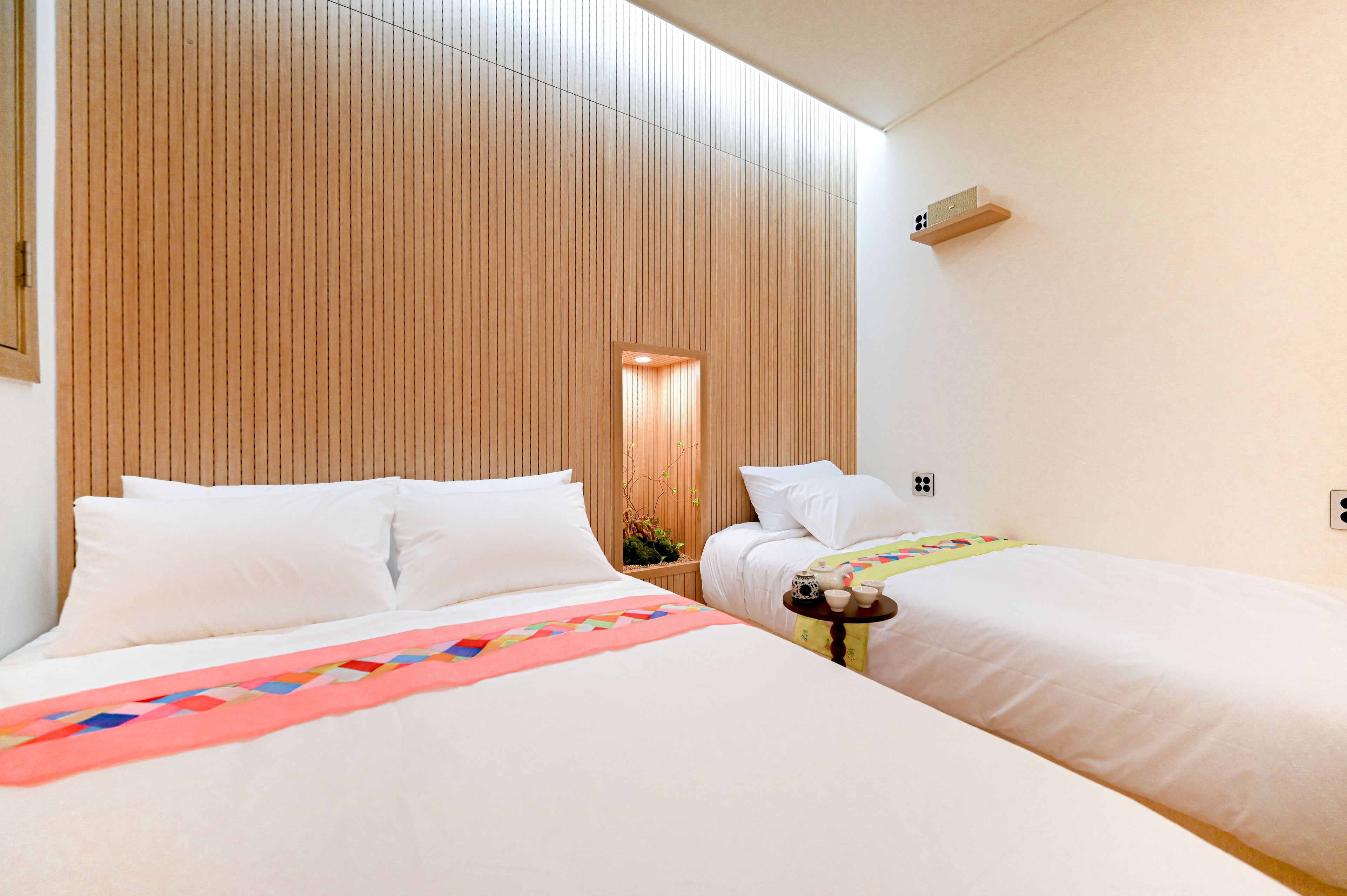 A hotel room with two beds, wooden paneling on the walls, a small table with a teapot and cups, and a wall-mounted air conditioning unit.