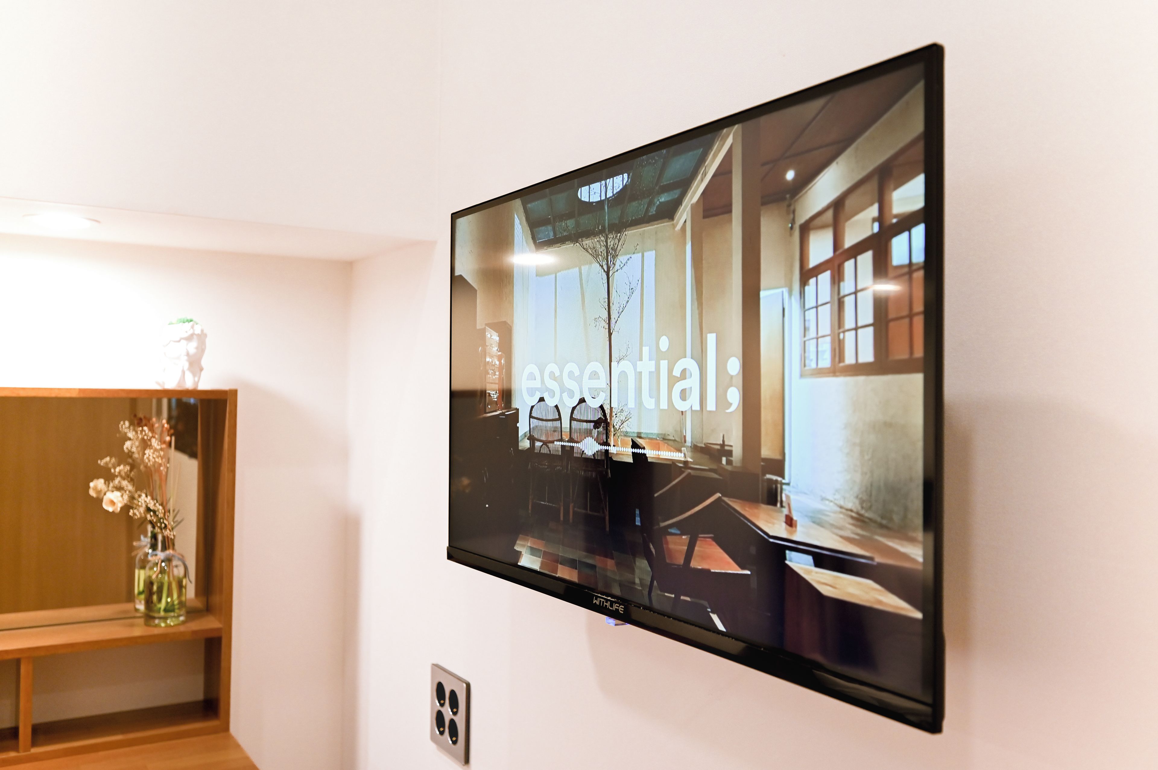 A flat-screen TV mounted on a wall displaying a room with a large window, a table, and chairs.
