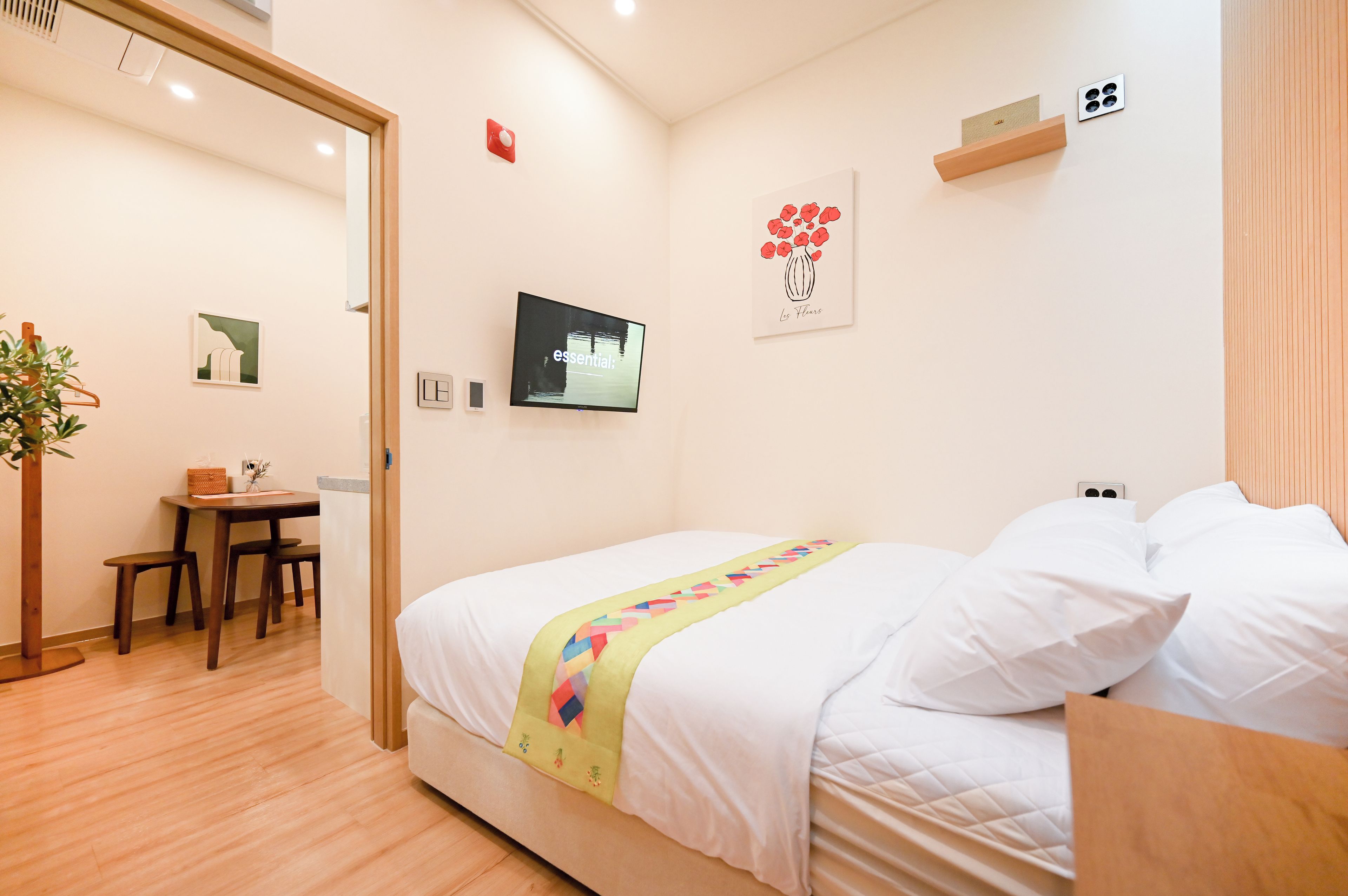 A hotel room with a bed, a desk, and a chair. There is a plant, a painting, and a wall-mounted television.