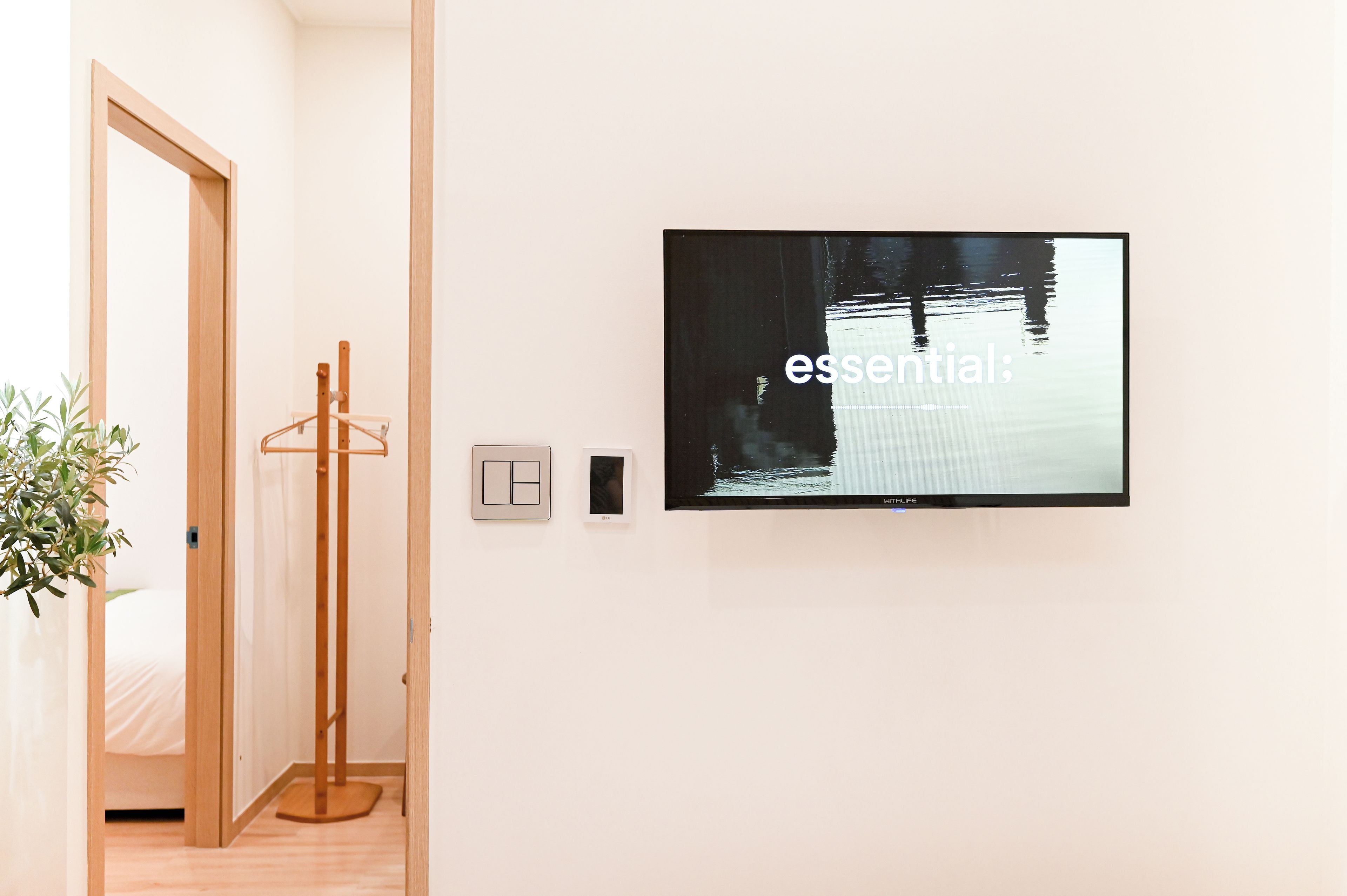 Quarto com televisão fixada na parede exibindo a palavra 'essential' e piso de madeira.