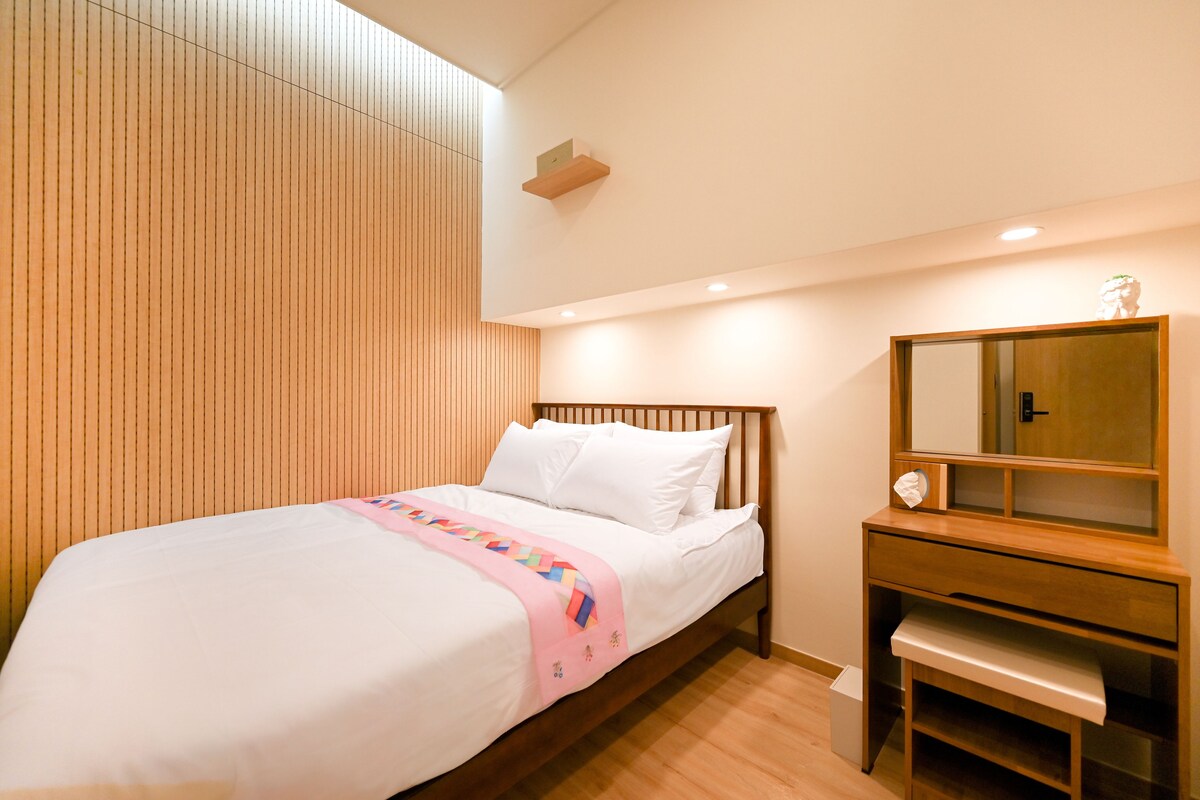 A hotel room with a wooden headboard, a bed with white bedding, a bedside table with a mirror, and a small shelf.