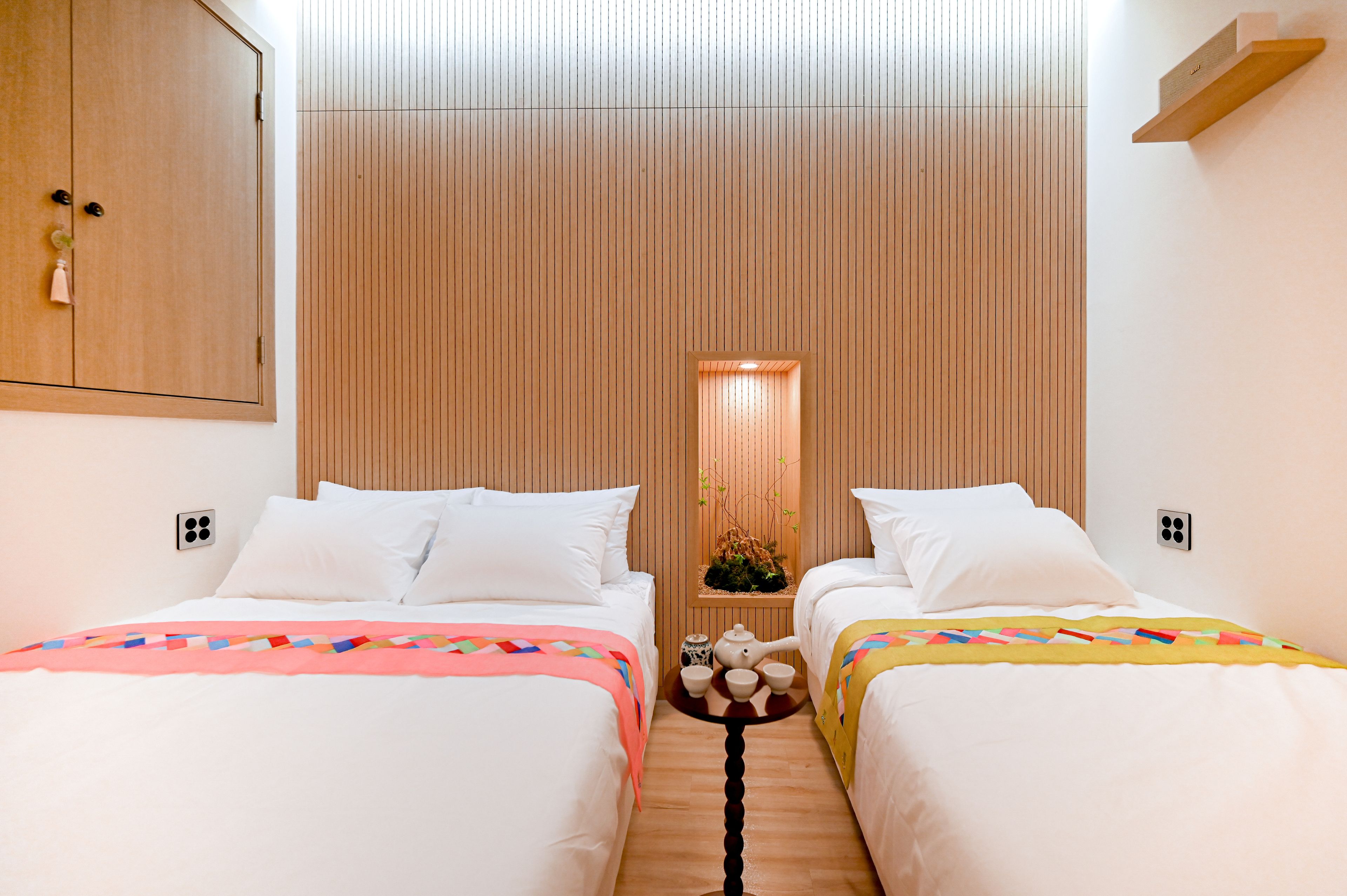 Habitación de hotel con dos camas, un cabecero de listones de madera, una mesita con tetera y una mesita de noche.