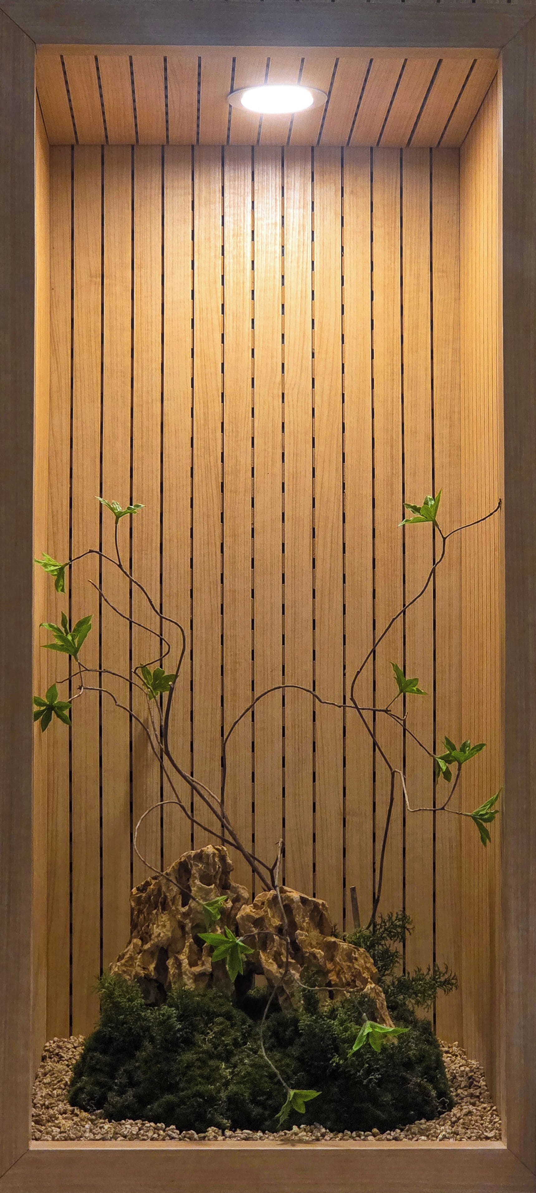 Pared vertical de madera con un hueco que muestra una formación rocosa, musgo y plantas pequeñas.