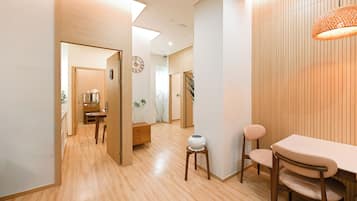 A modern interior with wooden flooring, a dining area with a table and chairs, a small stool with a vase, and a clock on the wall.