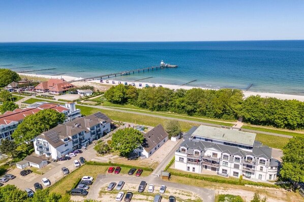 Eine Vogelperspektive eines Küstenhotels mit mehreren Gebäuden, einer Pier, die ins Meer hinausführt, und einem Parkplatz.