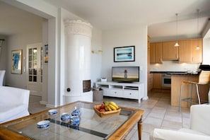 A modern living room with a glass coffee table, white sofas, and a kitchen area with wooden cabinets and a black countertop. - Holiday home with 1 bedroom (List auf Sylt)