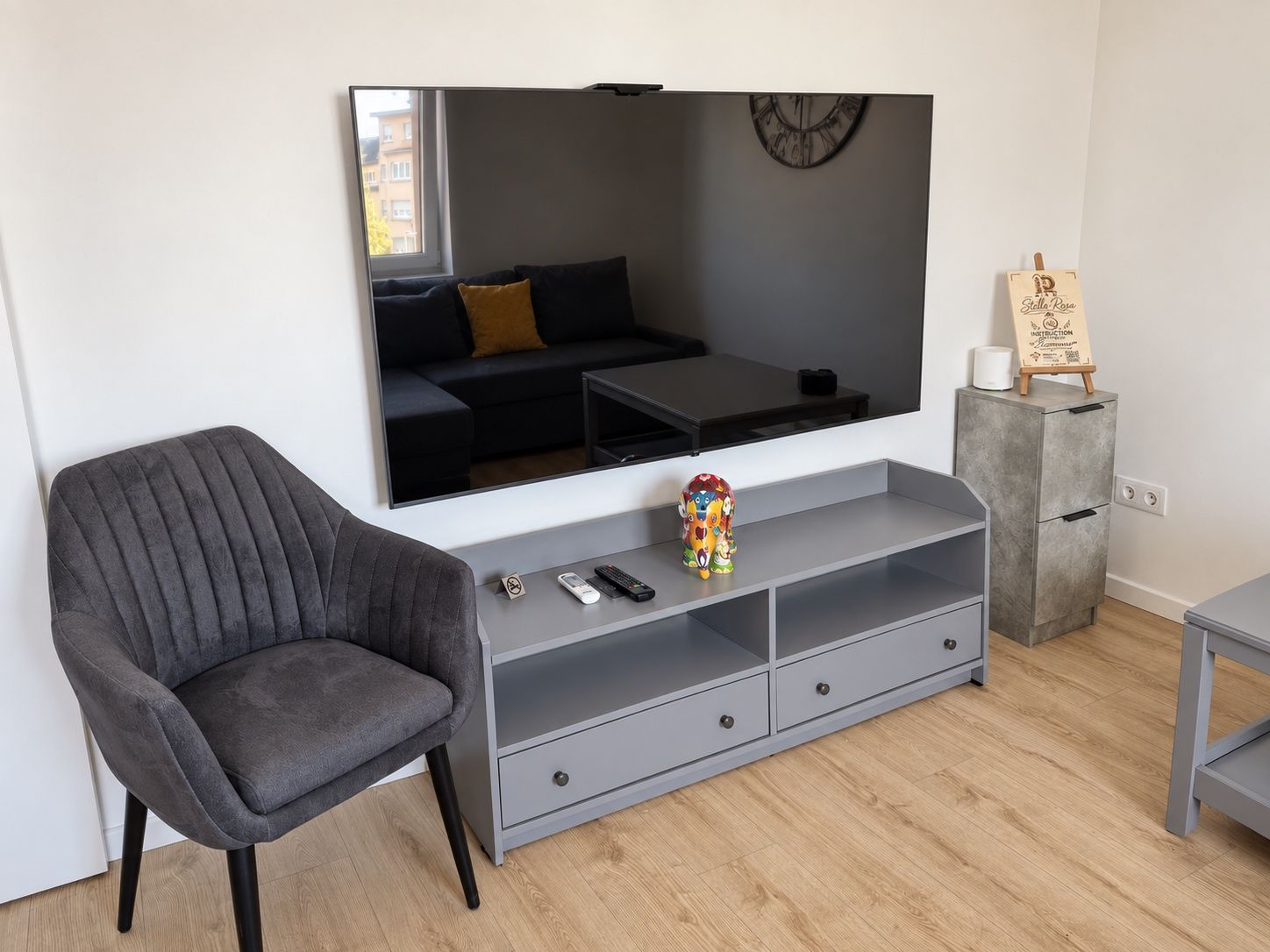 Uma sala de estar moderna com um sofá cinzento, uma televisão de tela plana grande, um console cinzento com gavetas, uma mesa pequena com um objeto decorativo e um armário cinzento com uma placa.