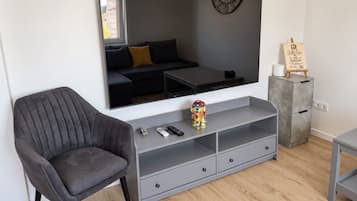 Uma sala de estar moderna com um sofá cinzento, uma televisão de tela plana grande, um console cinzento com gavetas, uma mesa pequena com um objeto decorativo e um armário cinzento com uma placa.