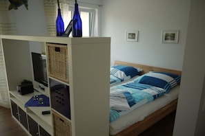 A bedroom with a bed, a television on a stand, and a shelf with items. - House of Four Elements (Zinnowitz)