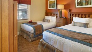 A hotel room with two beds, a nightstand, a lamp, and a window with blinds.