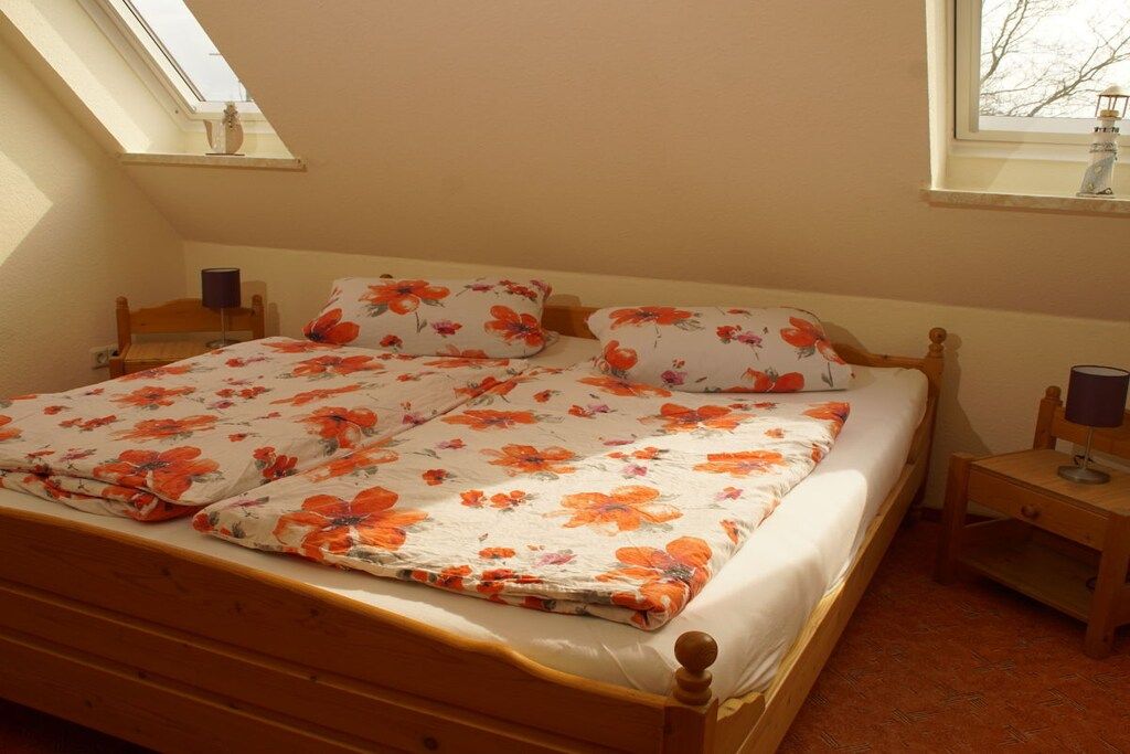 Una cama individual con cubrecama floral, cabecera de madera, mesita de noche con lámpara y ventana con vista a árboles.