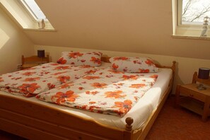 A single bed with a floral bedspread, a wooden headboard, a bedside table with a lamp, and a window with a view of trees.