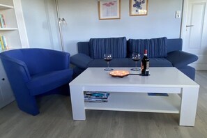 A living room with a blue sofa, a white coffee table, and two wine glasses. - Holiday apartment Hell (Wesselburener Deichhausen)
