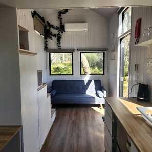Un espai habitable reduït amb sofà, màquina de cafè i finestra amb vista a vegetació.