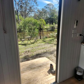 Une terrasse en bois avec une paire de bottes, une poubelle et une vue sur un paysage naturel composé d’arbres et d’une étendue d’eau.