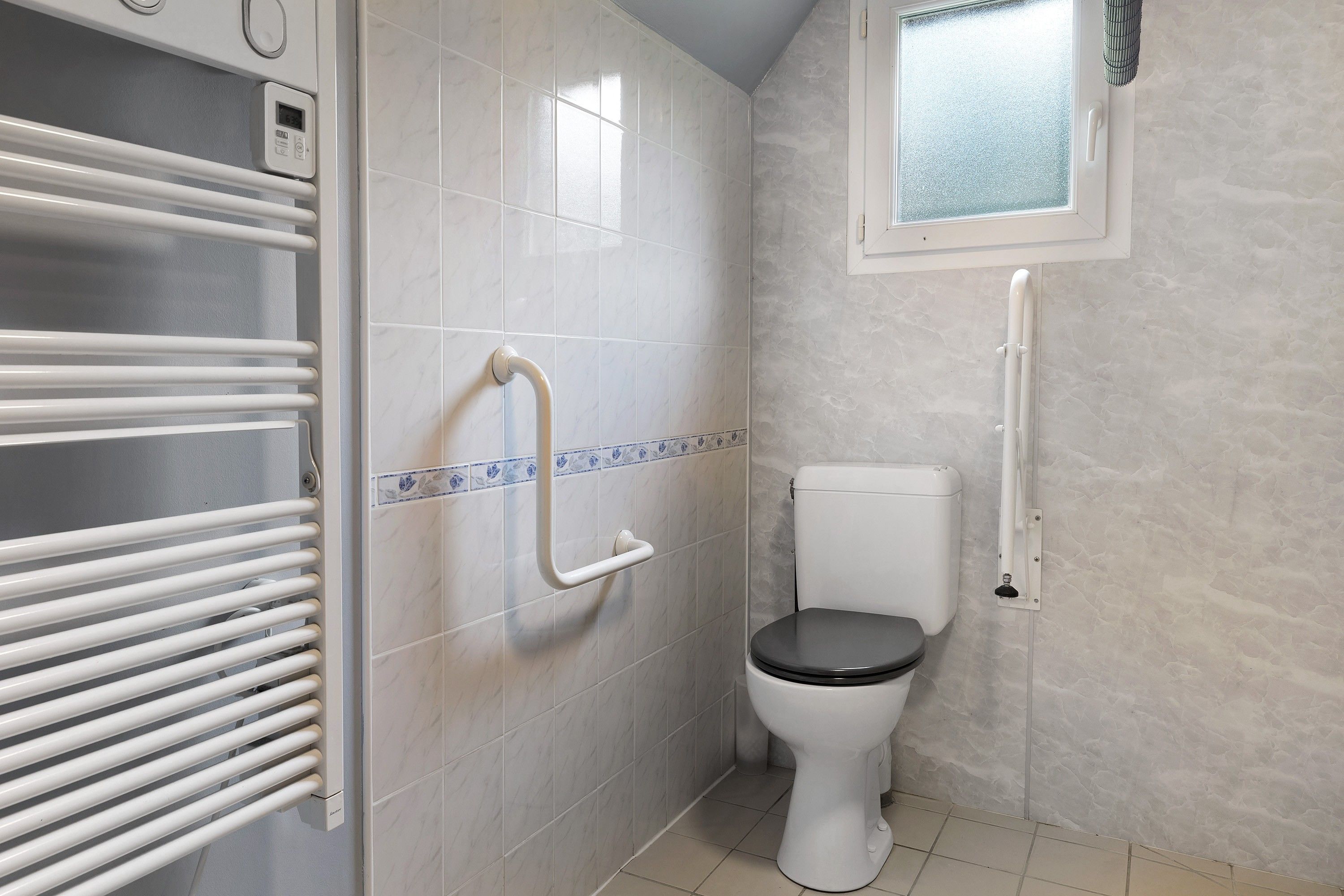 A bathroom with a toilet, a handrail, a towel rack, and a window.
