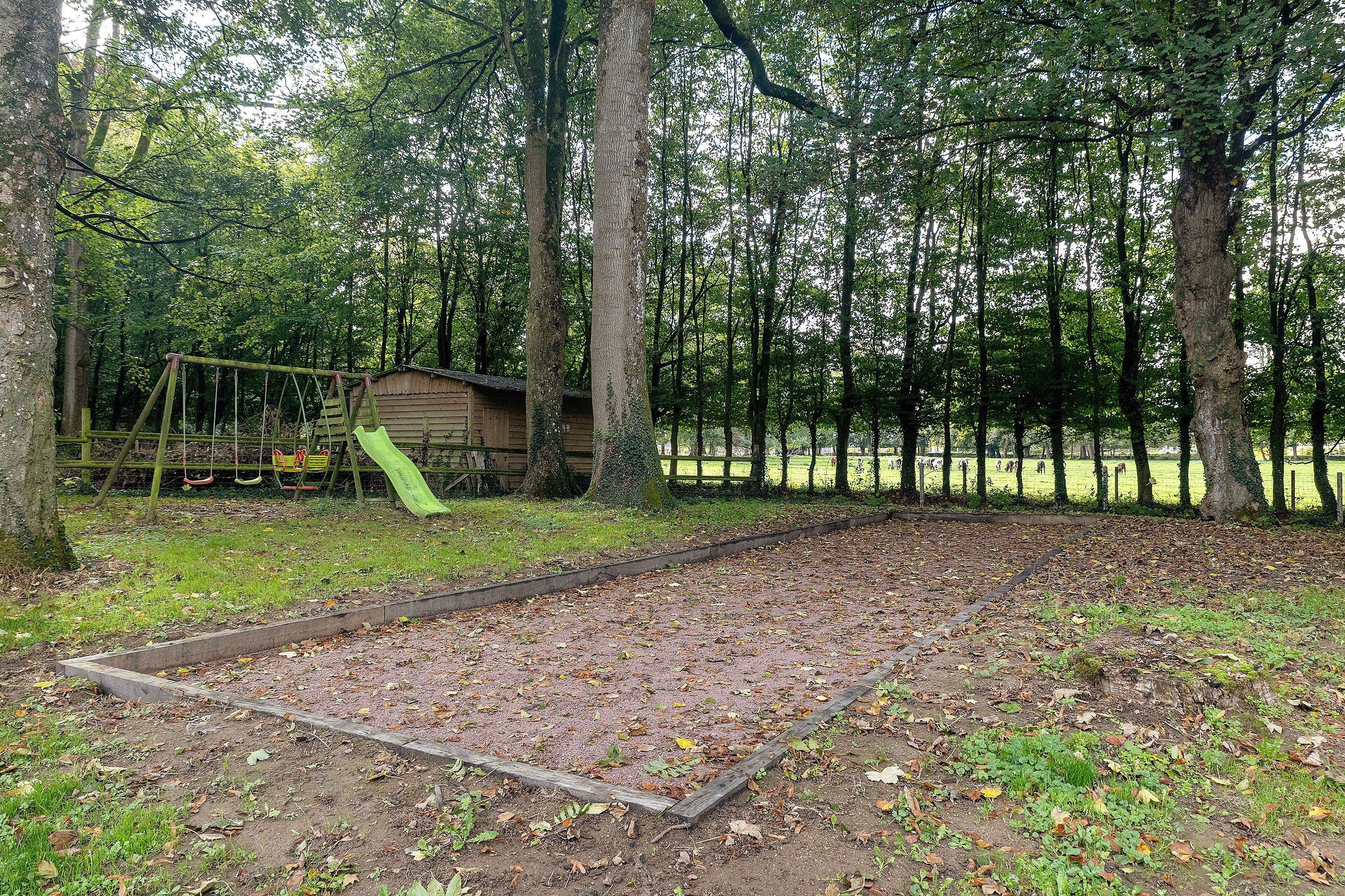 Een bebost gebied met een speeltuin, inclusief schommel en glijbaan, en een klein houten bouwwerk.