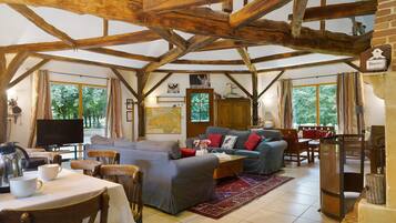 A rustic interior with exposed wooden beams, a dining area with a table and chairs, and a living room with sofas and a television.
