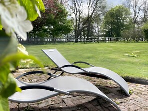 Deux fauteuils de jardin sont placés sur une terrasse en briques, avec une pelouse verdoyante et des arbres en arrière-plan.