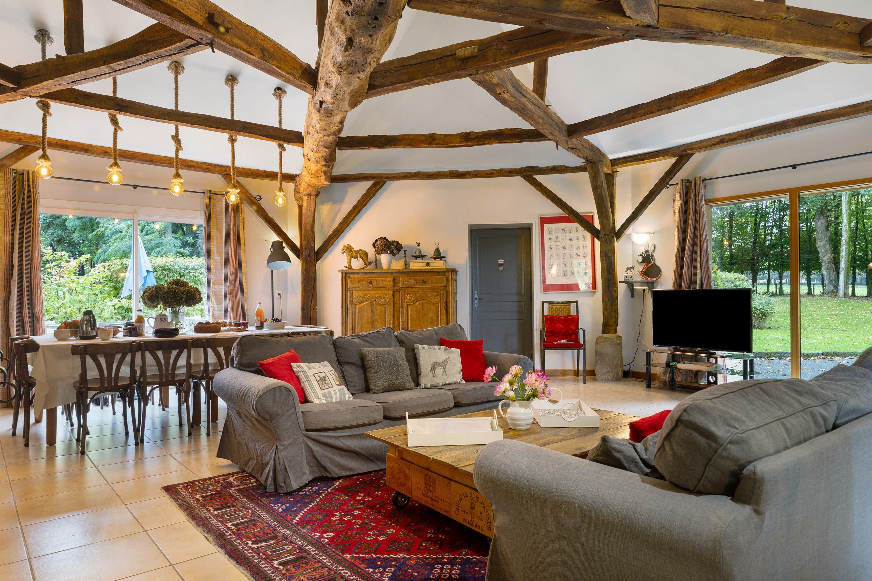 Een ruime woonkamer met een zithoek, een salontafel en een televisie. Er is een keukenhoek met een eettafel en stoelen. De kamer heeft zichtbare houten balken en grote ramen.