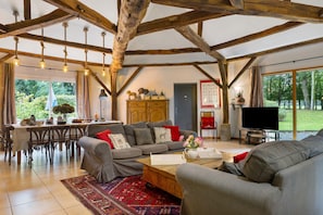 A spacious living area with a sofa set, a coffee table, and a television. There is a kitchen area with a dining table and chairs. The room has exposed wooden beams and large windows.