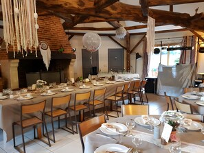 Une longue table à manger, entourée de chaises, une cheminée en briques et des décorations suspendues.