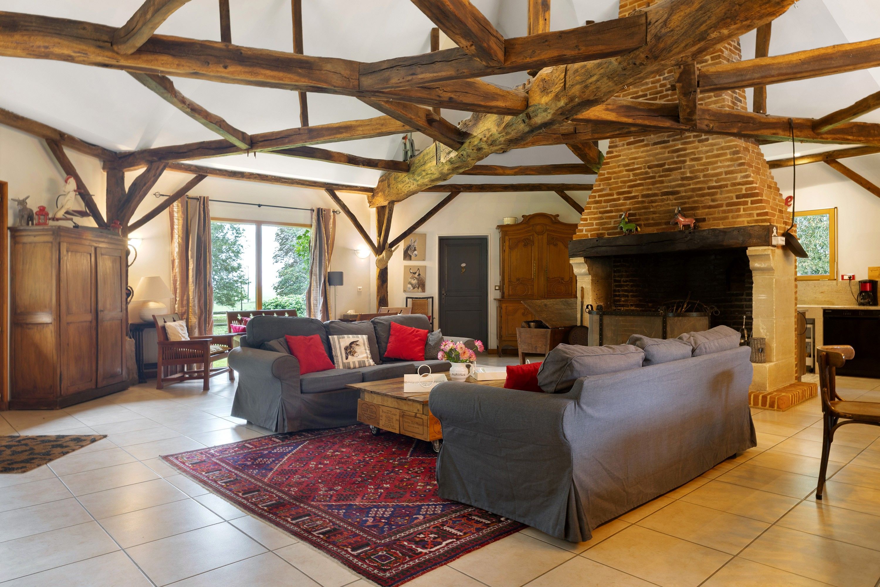 A spacious living room with a high ceiling, exposed wooden beams, a fireplace, and a large window providing natural light.