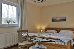 Un dormitorio con cama, una silla, una mesa con un florero, una ventana con vista a árboles y un cuadro en la pared.