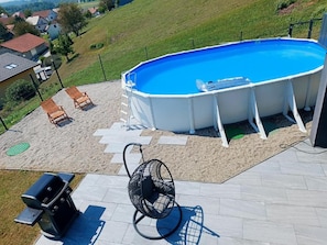 Ein Freibad mit klar blauem Wasser, umgeben von einer weißen Poolterrasse. Auf der Terrasse stehen zwei Holzliegestühle und ein schwarzer Eiersessel.