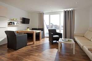 A modern living room with a sofa, a dining table with chairs, a flat-screen TV mounted on the wall, and a large window with curtains. - Winterstraße 17 Apartment 10 (Norderney)