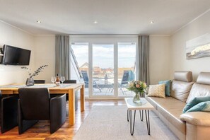 A modern living room with a dining area, a sofa, a TV mounted on the wall, and a view through large windows. - Winterstraße 17 Apartment 10 (Norderney)