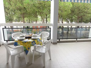 A dining area with a table set for four, white chairs, and a view of trees through a glass railing. - Sunny terrace apartment near beach (Bibione)